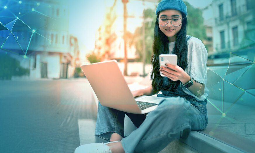 Junge asiatische Frau mit einer blauen Wollmütze und Laptop auf dem Schoß sitzt draußen in städtischem Umfeld und schaut auf ihr Smartphone